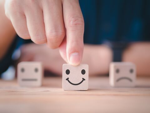 Hand placing a cube with a smiling face on a table, with two cubes showing neutral and unhappy faces in the background.